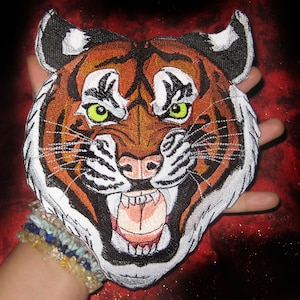 May include: A close-up of an embroidered tiger face patch. The tiger has orange and black stripes, white fur, and a fierce expression. The patch is perfect for adding a touch of wildness to clothing, bags, or other accessories.
