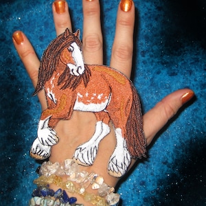 May include: An embroidered horse patch in brown and white, with a brown mane and tail. The patch is on a hand wearing bracelets.