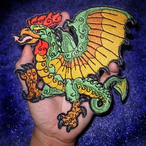 Puede incluir: Un parche bordado que representa una criatura mítica con cabeza de gallo, alas y cola larga, en tonos de verde, naranja y amarillo.