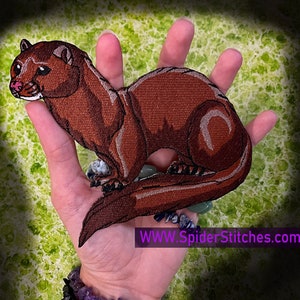 Puede incluir: Parche bordado marrón de una comadreja o un visón con el vientre blanco. El parche se sostiene en una mano con un fondo verde.