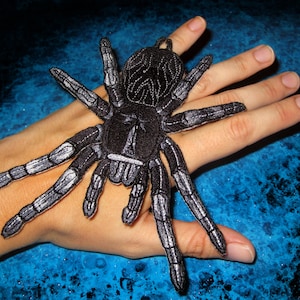 Könnte beinhalten: Schwarzer und silberner gestickter Vogelspinnen-Patch. Der Patch befindet sich auf einer Hand, die Beine der Vogelspinne sind ausgebreitet.