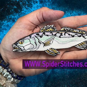 May include: A white embroidered patch of a fish with yellow and black details. The fish has a white body with black stripes and yellow fins. The patch is held in a hand with a bracelet on the wrist. The text "WWW.SpiderStitches.com" is visible below the patch.