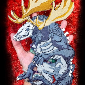 Puede incluir: Parche bordado que representa una criatura mítica con cabeza de calavera, cornamenta y cuerpo azul y gris. El parche tiene un diseño detallado con garras y dientes blancos. La dirección del sitio web WWW.SpiderStitches.com es visible en la parte inferior.