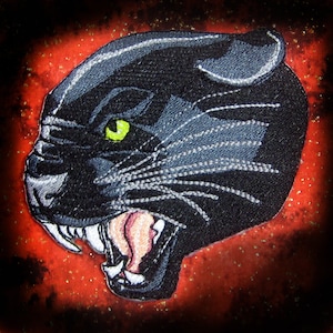 May include: A black panther with a green eye, snarling with its mouth open, showing sharp teeth. The panther is embroidered on a red fabric background.