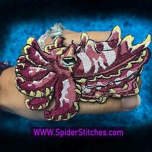 Op de afbeelding: Een geborduurde strijkpatch met een roze en gele sepia met een wit oog. De patch is op een hand met een paarse armband. De tekst "WWW.SpiderStitches.com" staat onderaan de afbeelding.