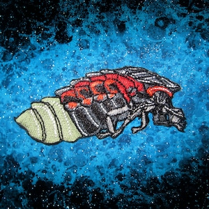 May include: A black and red embroidered insect with a green shell-like body against a blue and black speckled background.