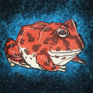 May include: Embroidered patch of a red and brown frog with white accents on a blue background with white speckles.