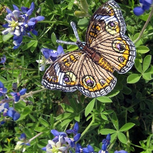 May include: A brown and yellow butterfly with white and purple accents is embroidered on a green fabric. The butterfly is surrounded by blue and white flowers.