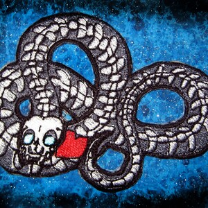 Puede incluir: Un parche bordado en blanco y negro de una serpiente con una calavera y un corazón rojo. La serpiente está enroscada en forma de ocho. El fondo es un cielo estrellado azul y blanco.