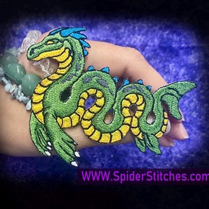 May include: Green and yellow embroidered patch of a sea serpent with blue and white details. The serpent is curled up with its tail in its mouth. The patch is on a hand.