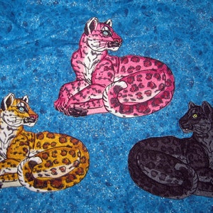 Puede incluir: Tres parches bordados de leopardos en diferentes colores. El primer leopardo es rosa con manchas negras, el segundo es marr&oacute;n con manchas negras y el tercero es negro con manchas grises.