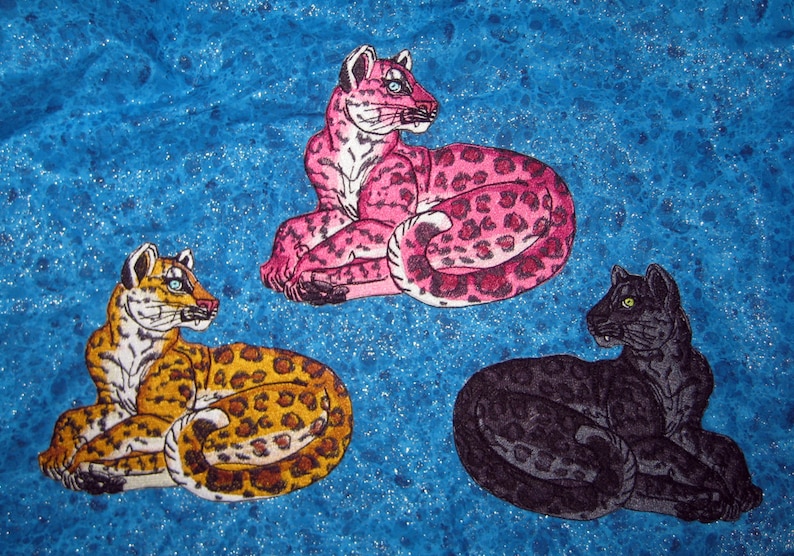 Puede incluir: Tres parches bordados de leopardos en diferentes colores. El primer leopardo es rosa con manchas negras, el segundo es marr&oacute;n con manchas negras y el tercero es negro con manchas grises.