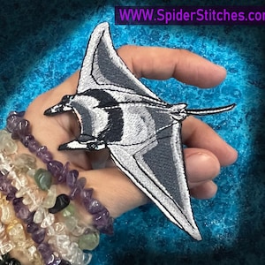 Pode incluir: Um remendo bordado cinza e branco de uma manta raia com contorno preto. O remendo é segurado em uma mão com várias pulseiras no pulso. O fundo é um design tipo aquarela azul e verde. O texto "WWW.SpiderStitches.com" está no canto superior direito da imagem.