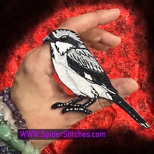Op de afbeelding: Zwarte en witte geborduurde vogelpatch met een lange staart. De patch wordt in een hand gehouden tegen een rode achtergrond. De tekst "WWW.SpiderStitches.com" is zichtbaar onderaan de afbeelding.