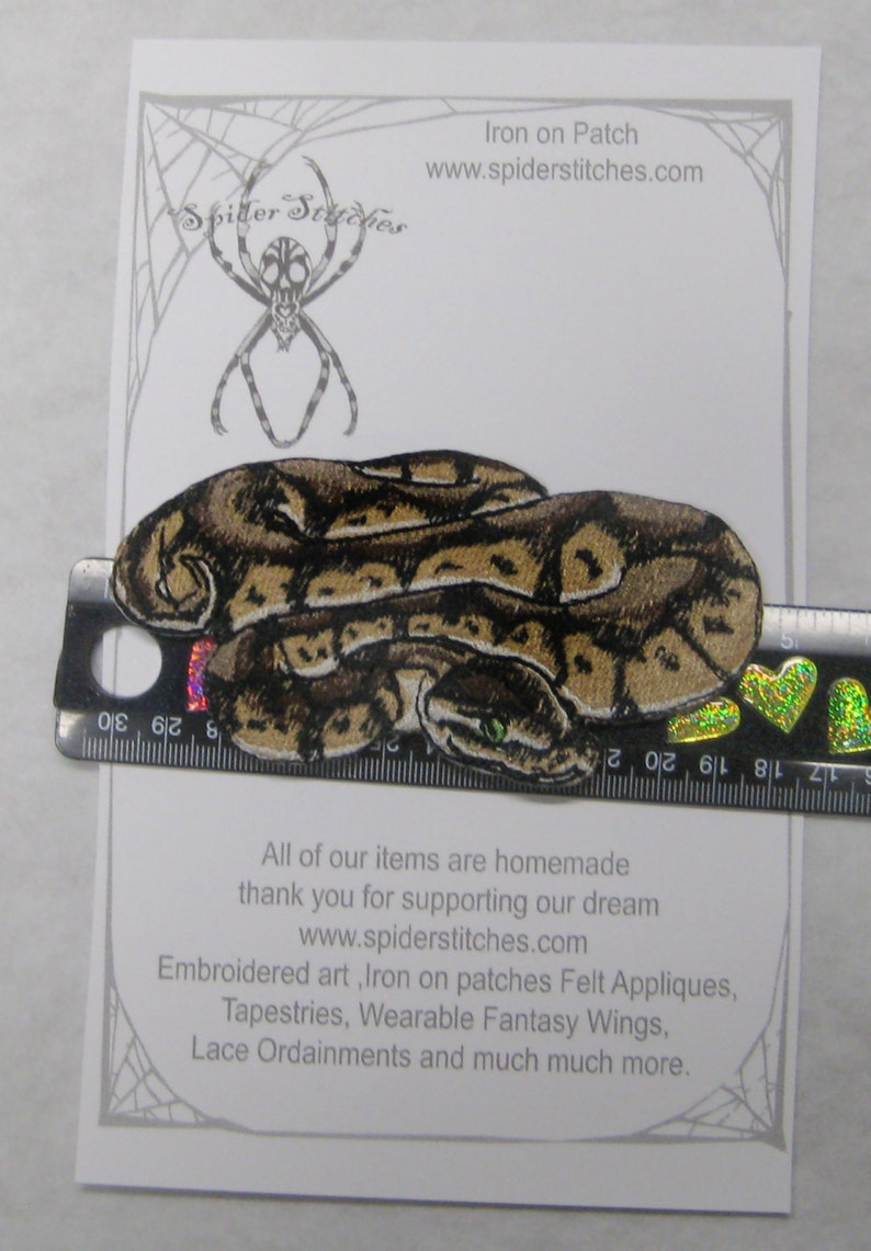 May include: Iron-on patch depicting a coiled brown and green snake with a textured surface. The patch measures approximately 7.5 cm in length. The text "Iron on Patch" and "www.spiderstitches.com" is printed on the white background.