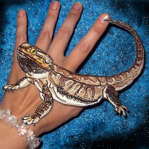 Peut inclure: Un patch brodé représentant un lézard barbu, avec des coutures brunes et blanches. Le patch est présenté sur la main d'une personne.