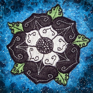 May include: Embroidered floral patch featuring a black and white flower with green leaves. The flower has a detailed design with a textured appearance. The background is a blue, speckled pattern.