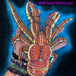 Op de afbeelding: Gestikt patch van een gele en oranje krab met zwarte ogen en poten. De krab bevindt zich op een blauwe achtergrond met de tekst "WWW.SpiderStitches.com" rechtsboven.
