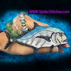 May include: Embroidered patch of a blue and white fish with a yellow eye. The fish is on a hand with a green and white bracelet.