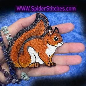 May include: Embroidered iron-on patch of a brown and white squirrel with a black bushy tail. The squirrel is sitting on its haunches with its tail curled up behind it.