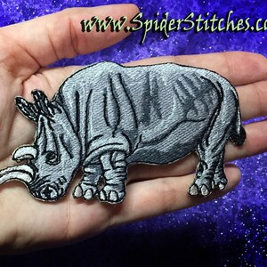 May include: A gray embroidered iron-on patch of a rhinoceros with a detailed design. The patch is held in a hand.