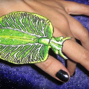 May include: A green and white embroidered ring featuring a detailed design of a sea creature. The ring is worn on a finger with black nail polish.