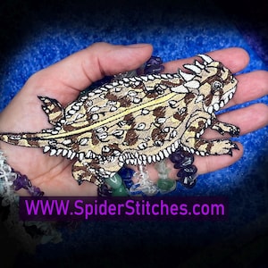 May include: Embroidered patch featuring a detailed depiction of a horned lizard. The patch showcases a brown and tan color palette with intricate stitching, including a yellow stripe down the back. The text "WWW.SpiderStitches.com" is visible.