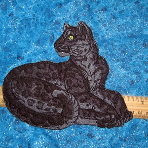 May include: Black embroidered patch of a panther with a green eye, lying down on a blue background with a ruler for scale.