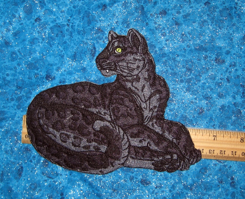 May include: Black embroidered patch of a panther with a green eye, lying down on a blue background with a ruler for scale.