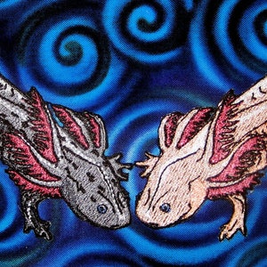 Puede incluir: Dos parches bordados de ajolote, uno con un cuerpo plateado y negro y el otro con un cuerpo marrón claro. Ambos ajolotes tienen branquias rojas plumosas y se miran.