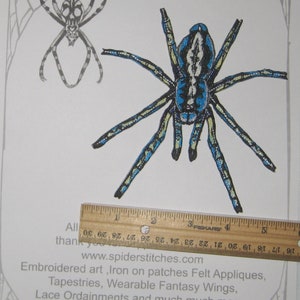 May include: Iron-on patch of a blue and black spider with yellow accents. The spider is approximately 4 inches wide. The patch is on a white background with a ruler for scale. The text "Iron on Patch" and "www.spiderstitches.com" is printed at the top of the image.