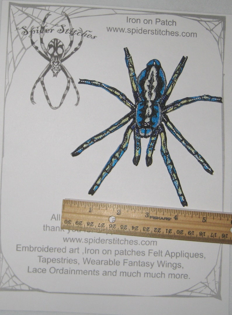 May include: Iron-on patch of a blue and black spider with yellow accents. The spider is approximately 4 inches wide. The patch is on a white background with a ruler for scale. The text "Iron on Patch" and "www.spiderstitches.com" is printed at the top of the image.