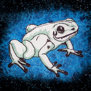 May include: A white frog with black eyes and a black outline is embroidered on a blue fabric with white speckles. The frog is facing the right side of the image.