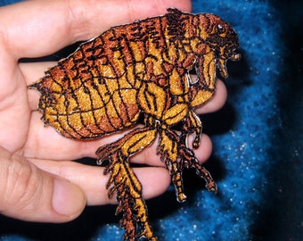 May include: A detailed embroidered patch of a flea, with a brown and gold color scheme. The flea is depicted in a realistic style, with intricate details on its body and legs.