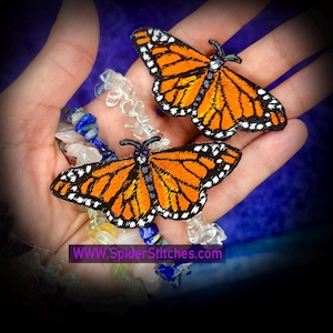 May include: Two embroidered monarch butterfly patches featuring vibrant orange wings with black veins and white-spotted black borders. The patches are displayed alongside a multi-colored gemstone chip bracelet. The text WWW.SpiderStitches.com is visible.