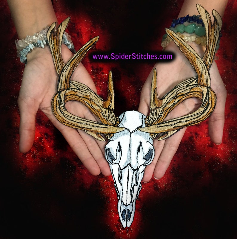 May include: A white deer skull with large, intricately embroidered antlers in shades of brown and gold. The skull is held in the palms of two hands, with the text "www.SpiderStitches.com" visible at the top of the image.