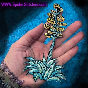 May include: An embroidered iron-on patch depicting a desert plant with a tall stalk of yellow flowers. The patch is blue and yellow and is shown on a hand, with the website "WWW.SpiderStitches.com" visible in the background.