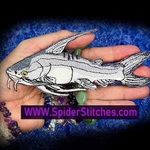 May include: An embroidered patch shaped like a catfish with a white body, grey shading, and a yellow eye. The patch features detailed stitching for the fins, tail, and whiskers. The text WWW.SpiderStitches.com is printed in purple at the bottom.