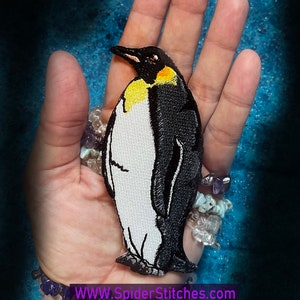 May include: A black and white embroidered penguin patch with a yellow chest. The patch is on a hand with several bracelets.