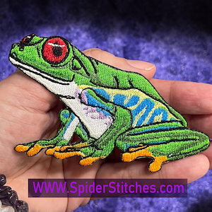 Pode incluir: Etiqueta bordada verde e azul de uma rã-árvore de olhos vermelhos. A rã está sentada sobre as suas patas traseiras com as patas dianteiras estendidas. A etiqueta tem aproximadamente 7,5 cm de comprimento.