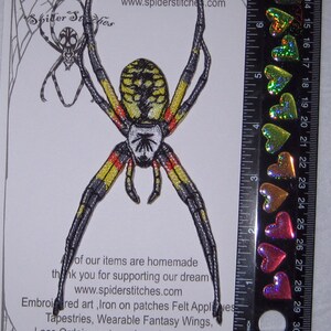 May include: Iron-on patch of a yellow and black spider with red markings. The spider is approximately 7.5 cm tall. The patch is on a white background with a spider web design. The text "Iron on Patch" and "www.spiderstitches.com" is printed on the background.