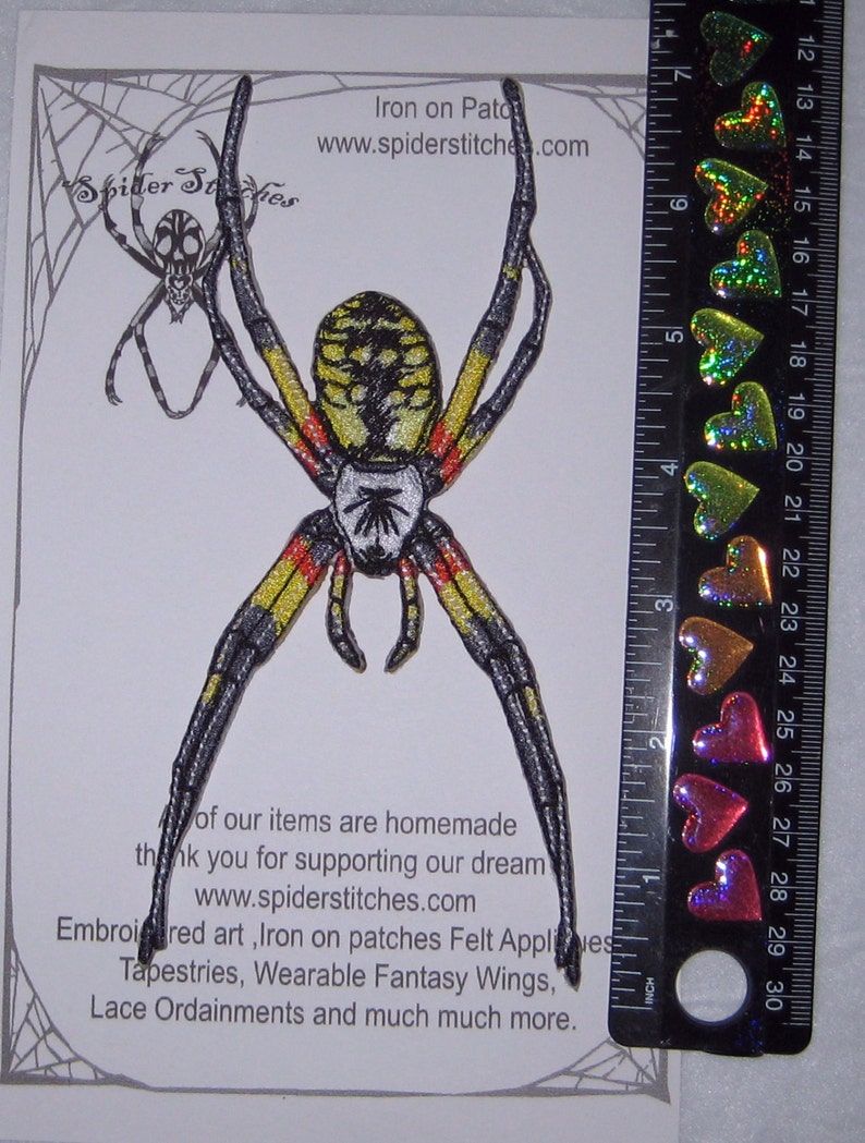 May include: Iron-on patch of a yellow and black spider with red markings. The spider is approximately 7.5 cm tall. The patch is on a white background with a spider web design. The text "Iron on Patch" and "www.spiderstitches.com" is printed on the background.