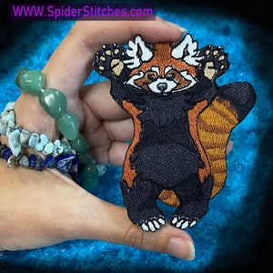 Peut inclure: Un patch brodé de panda roux avec un corps noir, des pattes orange et un visage blanc. Le patch lève les pattes en l'air. Le patch mesure environ 10 cm de haut.