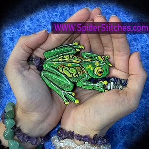 May include: Green embroidered frog patch with white stitching and yellow accents. The patch is held in the palm of a hand with a purple and green gemstone bracelet on the wrist.