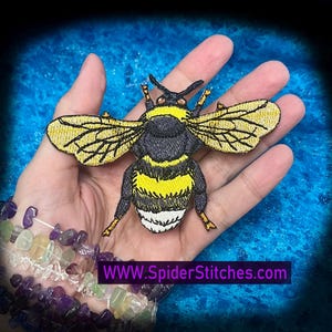 May include: Embroidered bumblebee patch with yellow and black stripes, detailed wings, and the text WWW.SpiderStitches.com. The patch is held in a hand, with a beaded bracelet in the foreground.