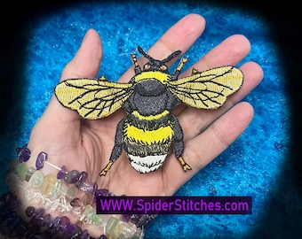 Peut inclure: Écusson brodé d'un bourdon avec des rayures jaunes et noires, des ailes détaillées et le texte WWW.SpiderStitches.com. L'écusson est tenu dans une main, avec un bracelet de perles au premier plan.
