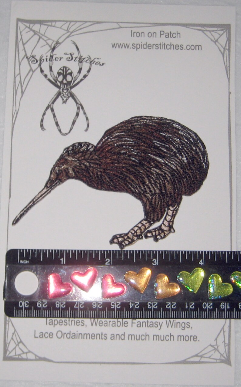 Op de afbeelding: Een opstrijkbare patch met een gedetailleerde illustratie van een kiwi-vogel. De patch is bruin met een lange snavel en is ongeveer 7.6 cm lang. De afbeelding bevat ook hartvormige versieringen en de tekst "Spider Stitches".