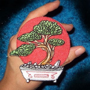 Puede incluir: Parche bordado con un árbol bonsái en una bañera blanca sobre un fondo rojo. El árbol tiene hojas verdes y un tronco marrón. La bañera está llena de piedras grises.