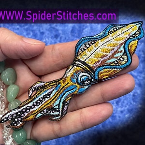 Puede incluir: Un parche bordado de hierro de un calamar en azul, dorado y blanco. El parche está en una mano con un brazalete de cuentas.