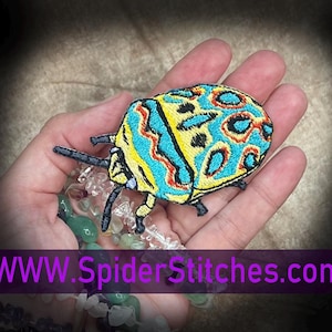 Puede incluir: Parche bordado de insecto con un diseño colorido en amarillo, azul y rojo. El parche tiene un patrón de insecto detallado y se sostiene en una mano. El texto "WWW.SpiderStitches.com" se muestra en morado.